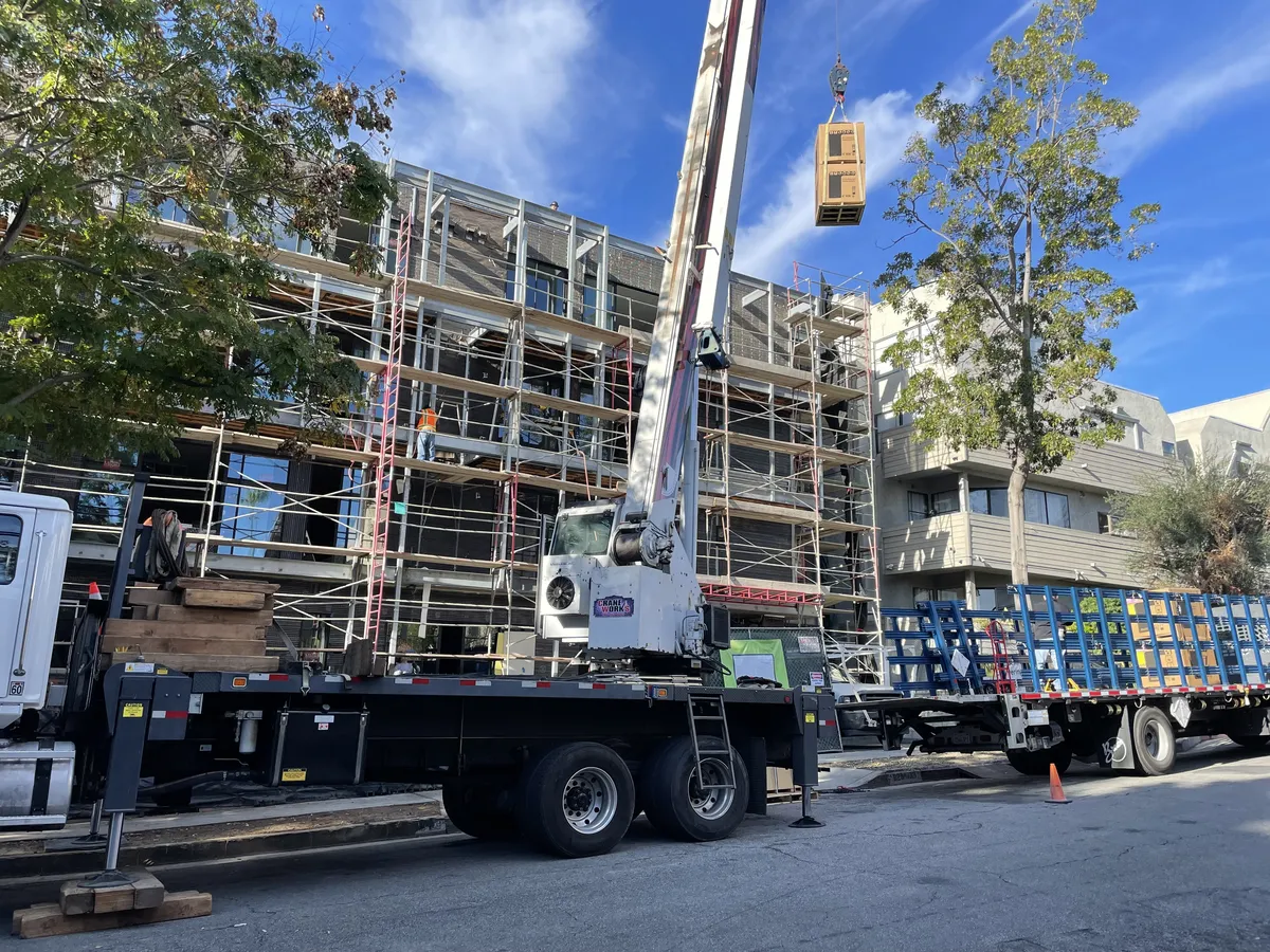 Crane truck lifting a load beside a multistory building wrapped in scaffolding.