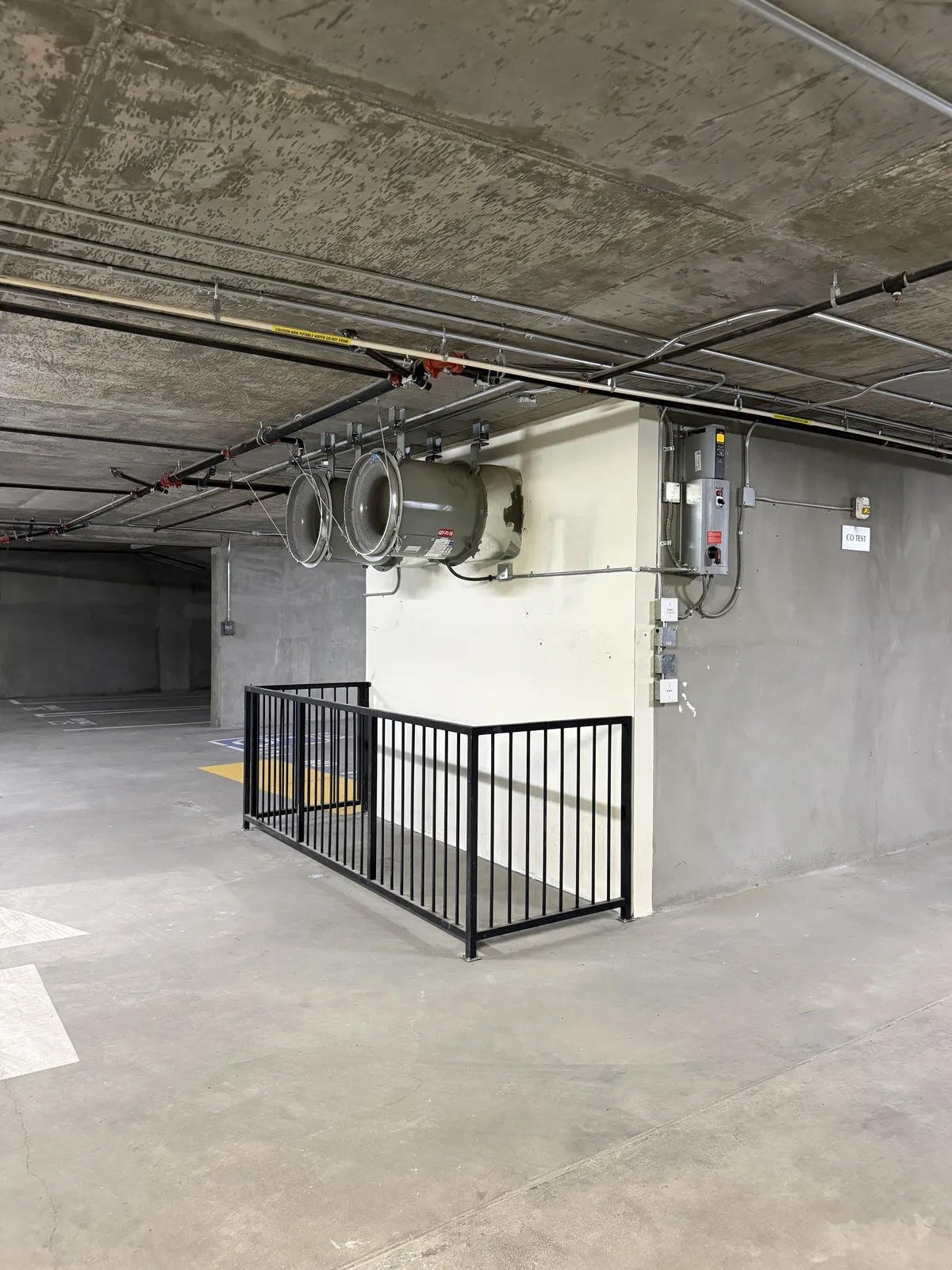 Two ventilation fans mounted high on a concrete wall above a black railing in a parking garage.