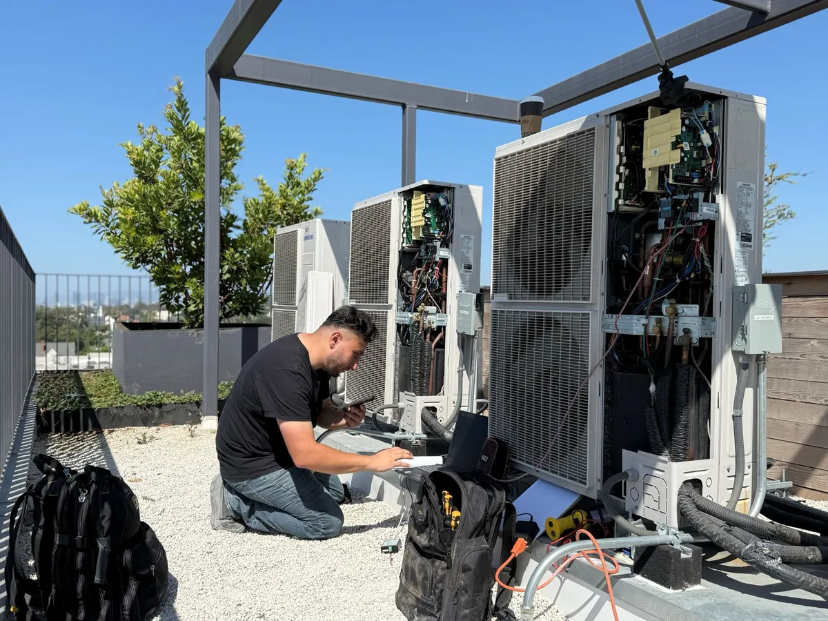 Air Plus HVAC technician kneeling beside open outdoor units on a rooftop.