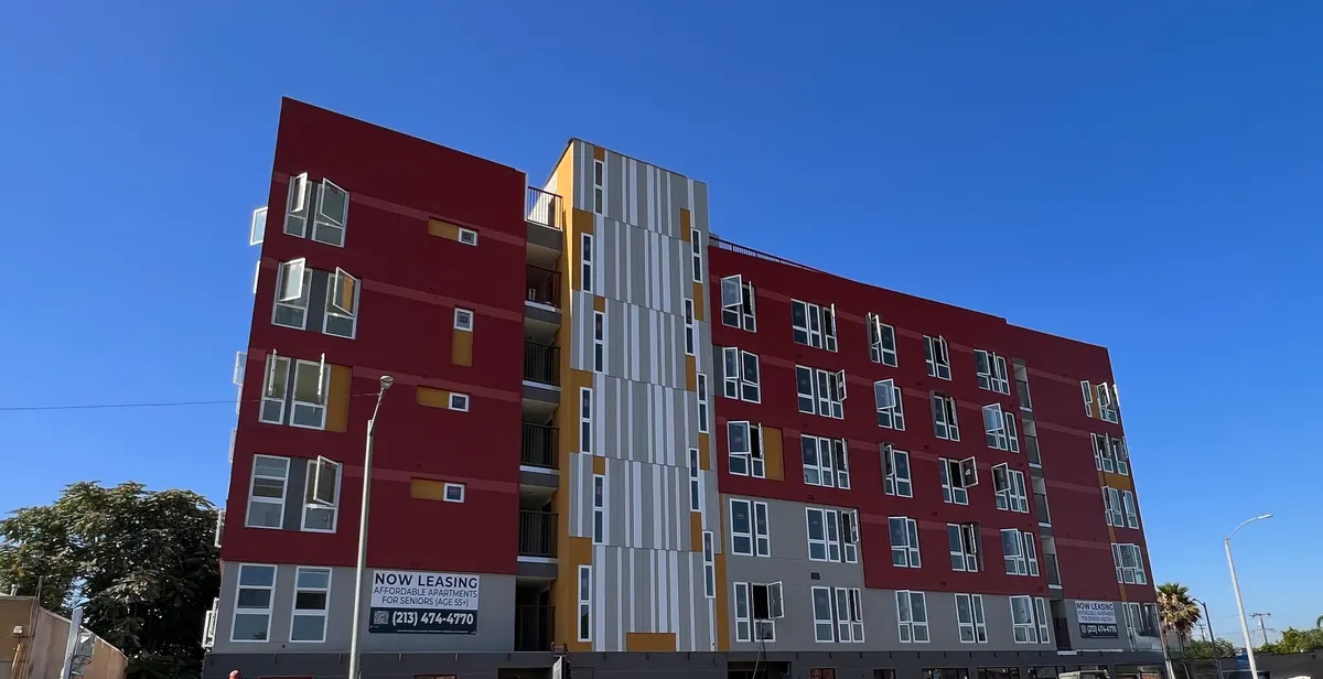 Six-story apartment building with a red and gray facade against a clear blue sky.