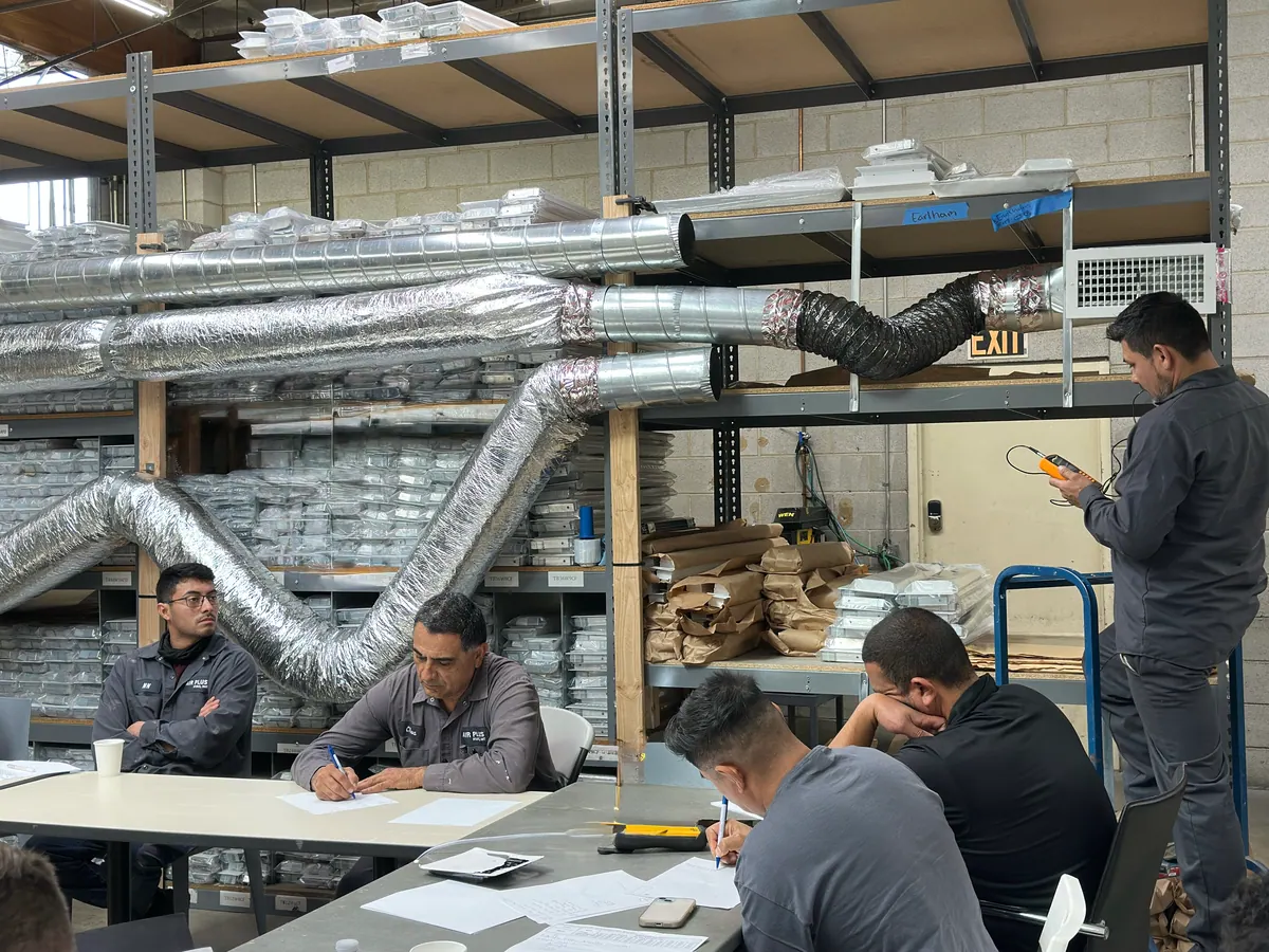 Air Plus HVAC crew members around a table in the shop, with ductwork racked overhead.