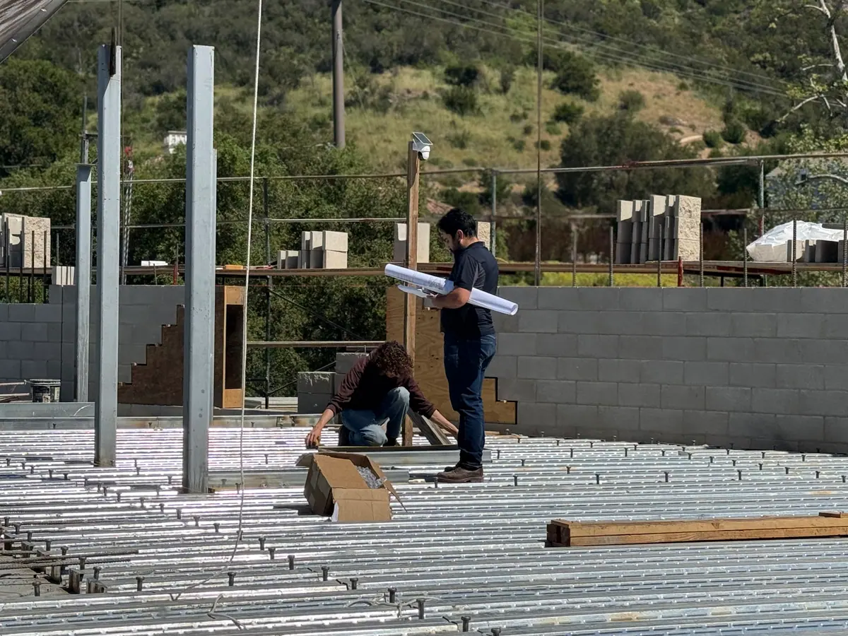 Two Air Plus HVAC crew members on a metal floor deck at a construction site, one holding rolled plans.