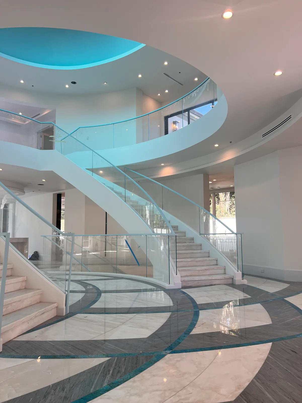 Curved glass staircase with a patterned marble floor and a blue-lit ceiling.