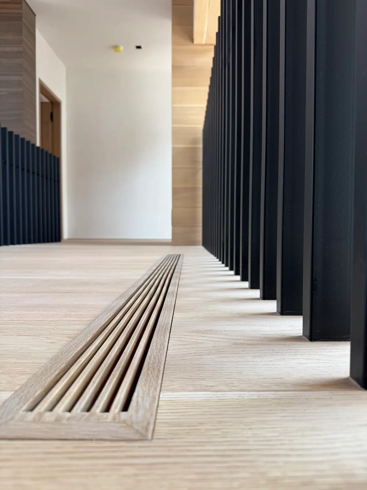 Linear floor diffuser set flush into a light oak floor beside a black railing.