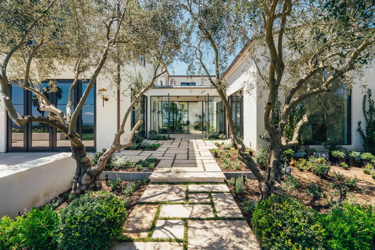 Stone-paved path through an olive-tree courtyard to the glass entry doors of a white home.