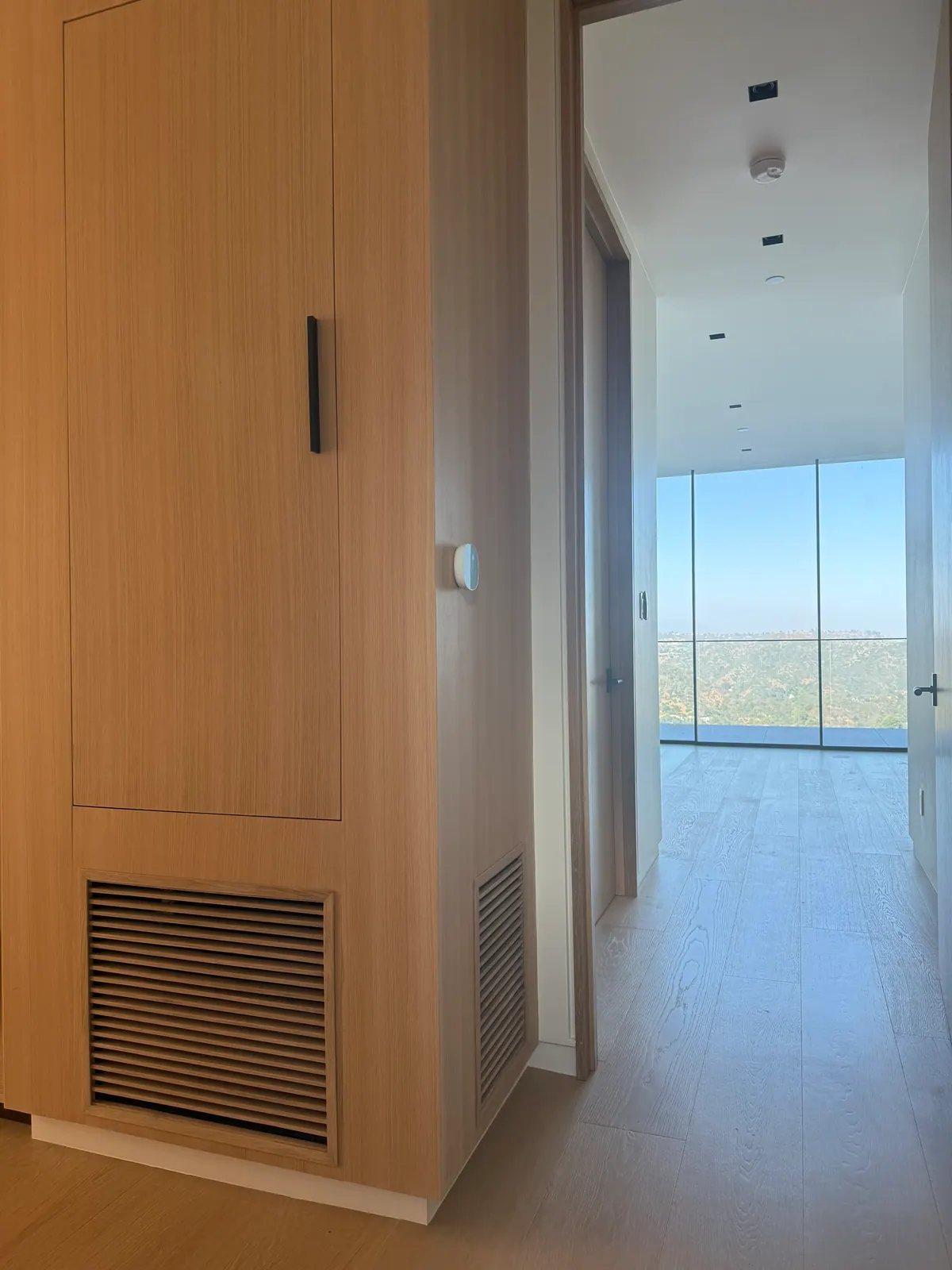 Louvered grille at the base of a tall oak panel wall, with a thermostat beside it.