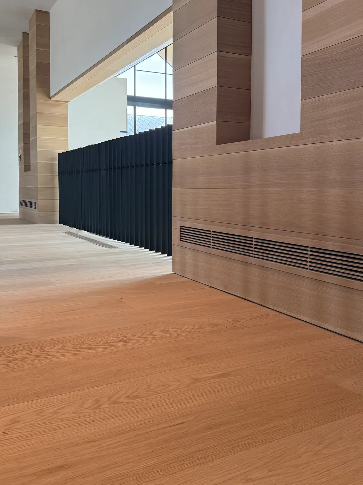 Long slot vent at the base of a wood-paneled wall above an oak floor.