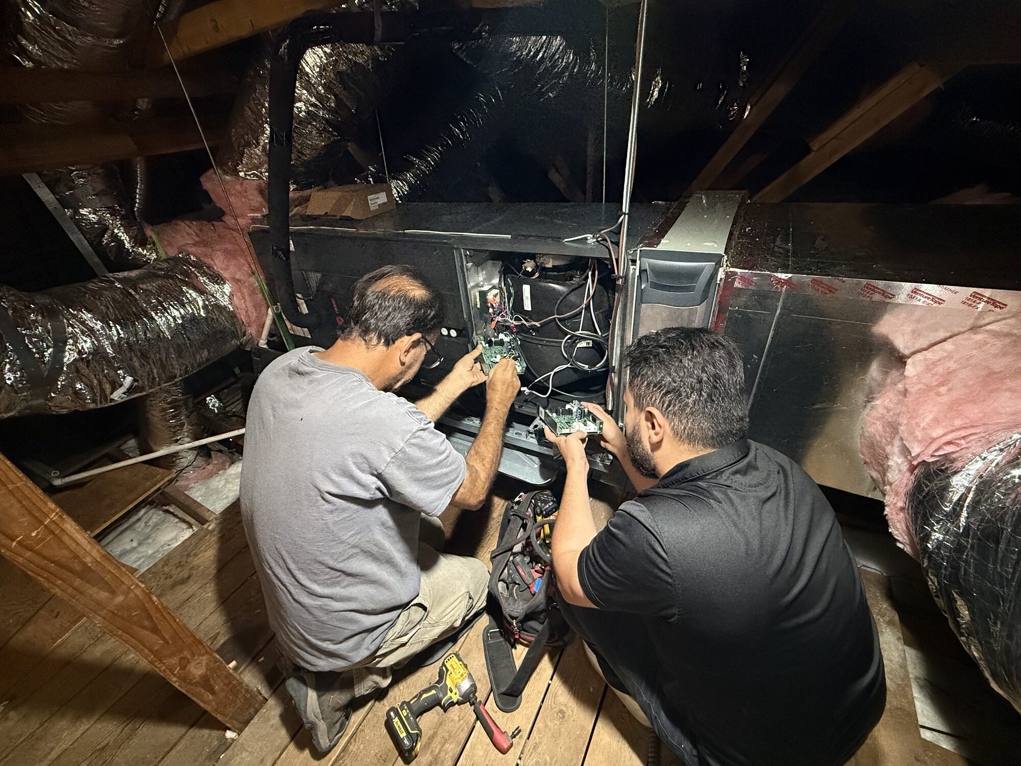 Two technicians working on a furnace control board in an attic.