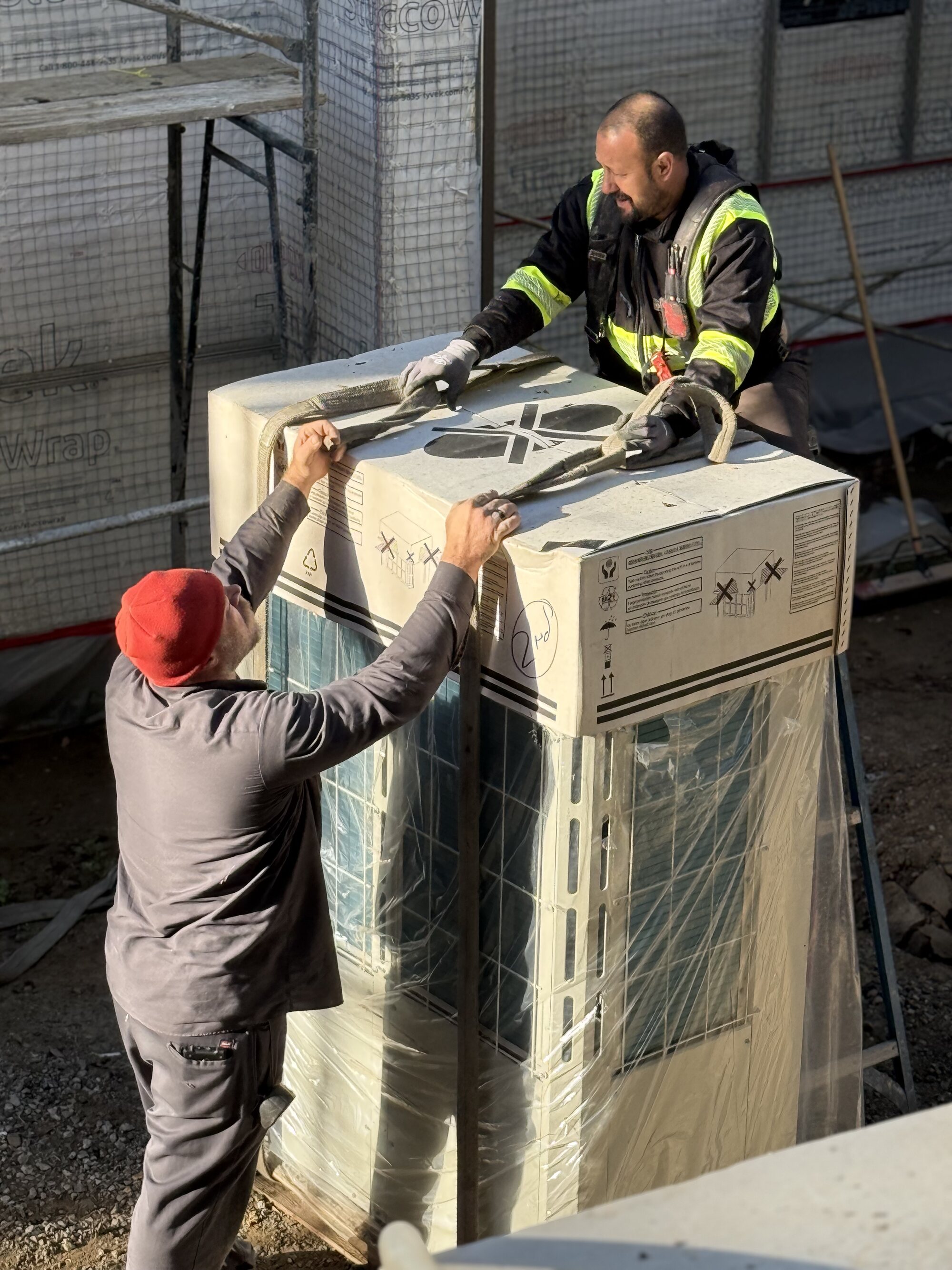 Two crew members rigging a boxed outdoor unit on a construction site.