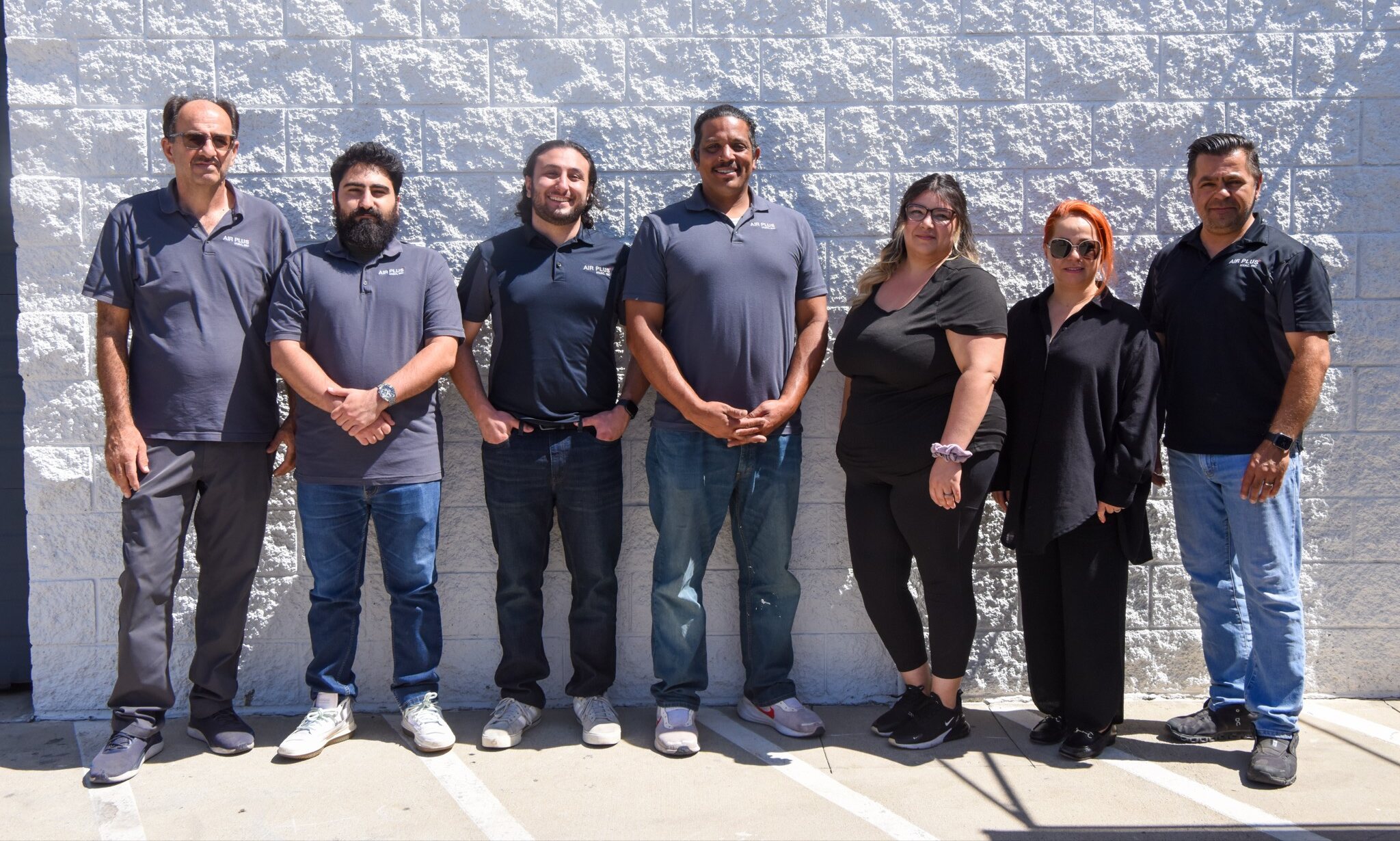 Seven members of the Air Plus HVAC headquarters team standing together in front of a white block wall.