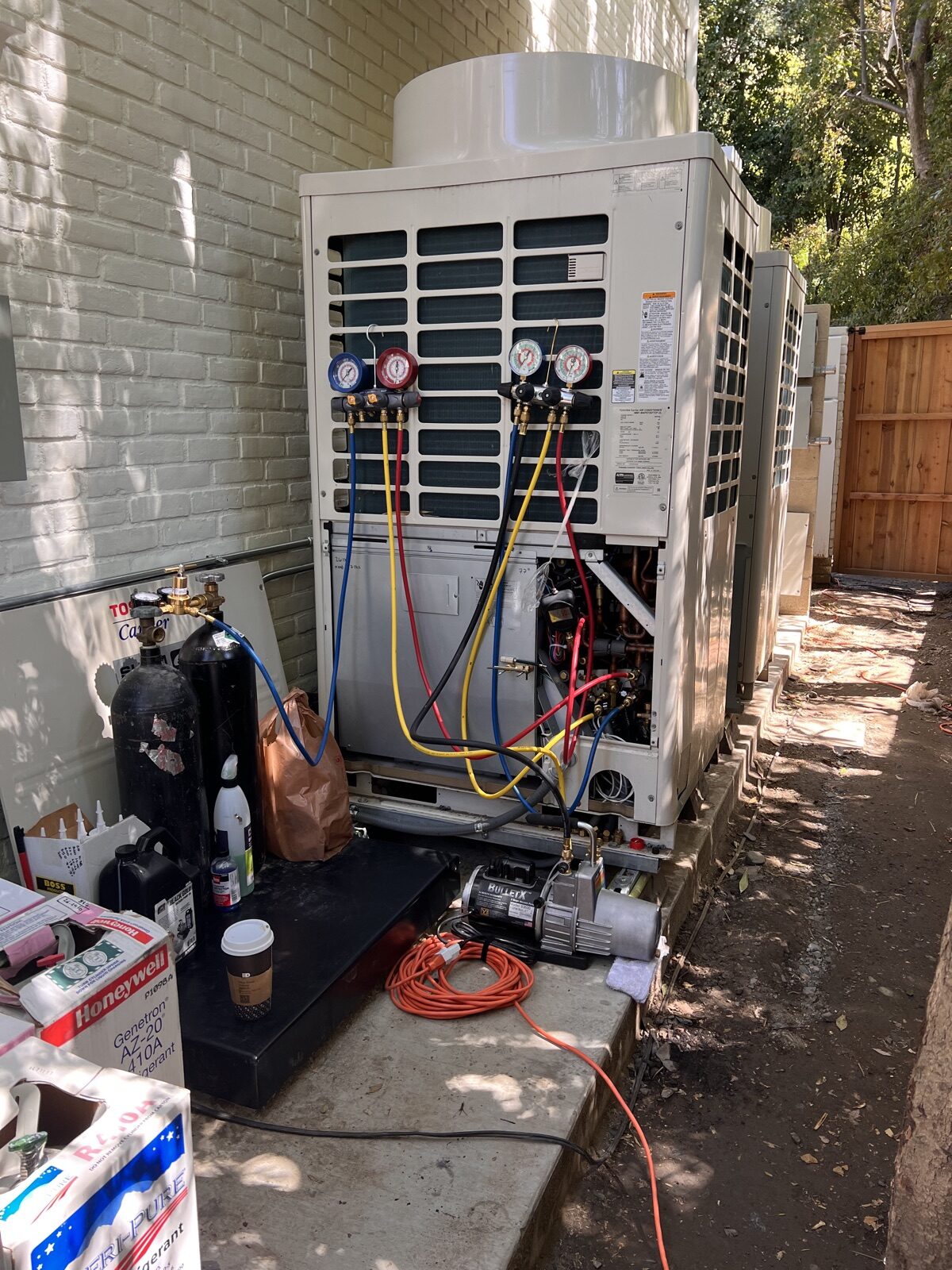 Refrigerant gauges connected to an outdoor unit in a side yard, with a vacuum pump on the ground.