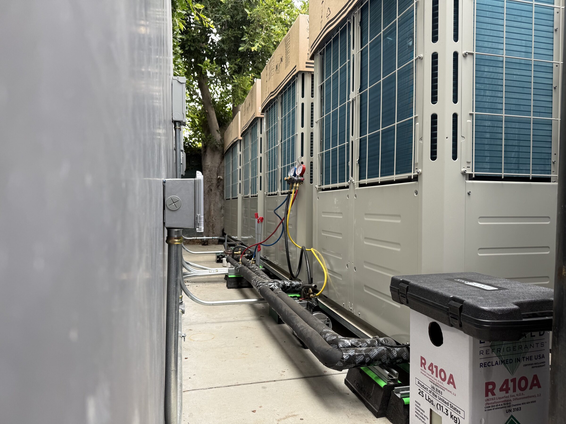 Large outdoor units along a side wall with refrigerant gauges connected and a refrigerant tank beside them.