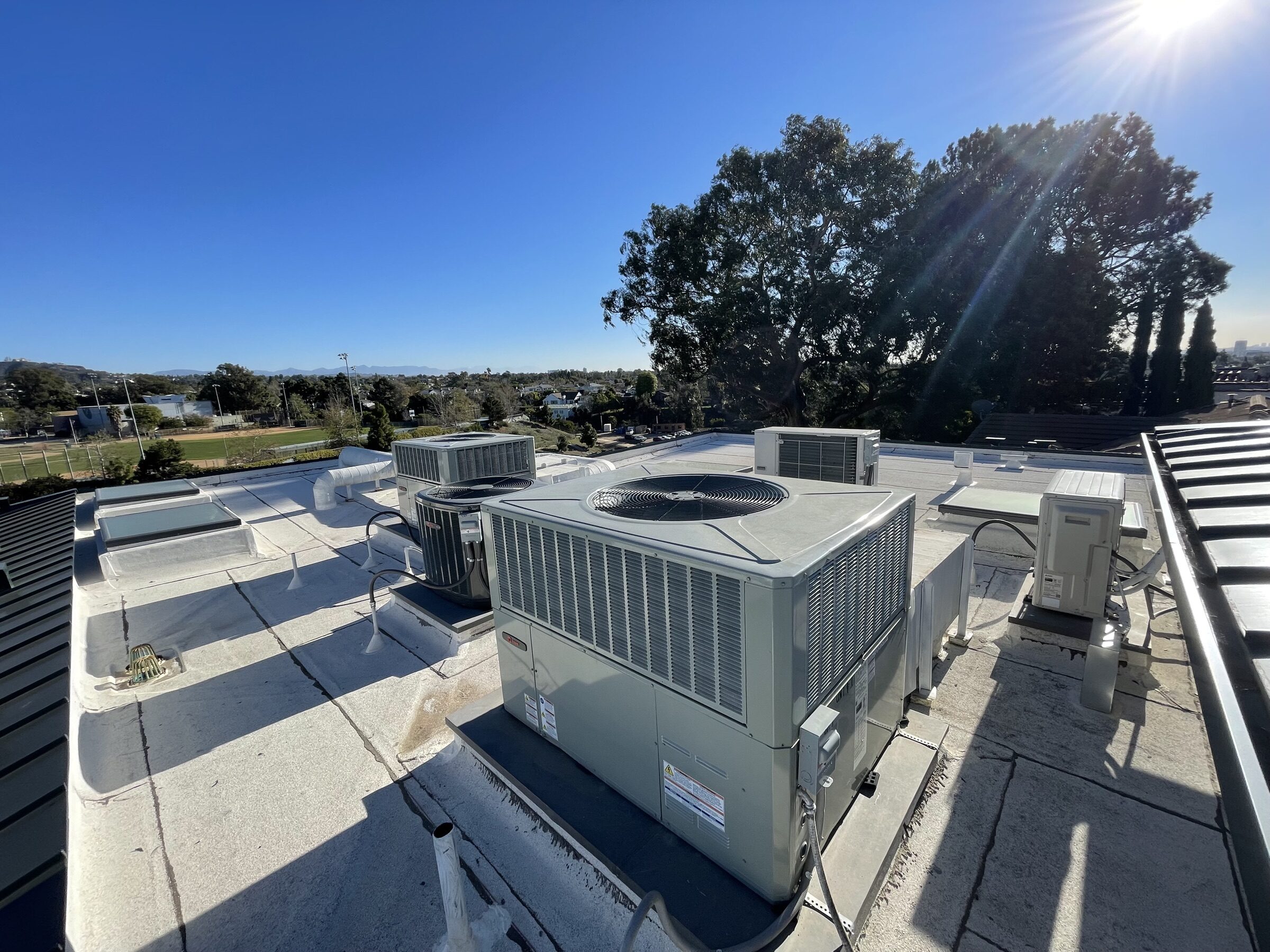 A rooftop air conditioning unit in strong sun, with trees and a blue sky behind it.
