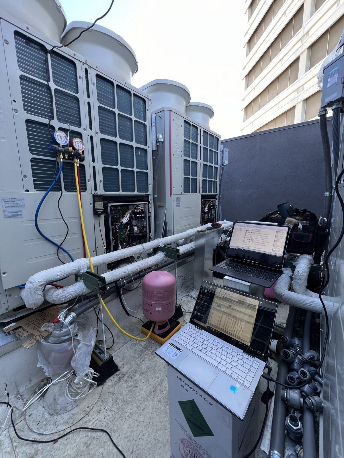 Refrigerant gauges and a laptop connected to outdoor units on a rooftop.