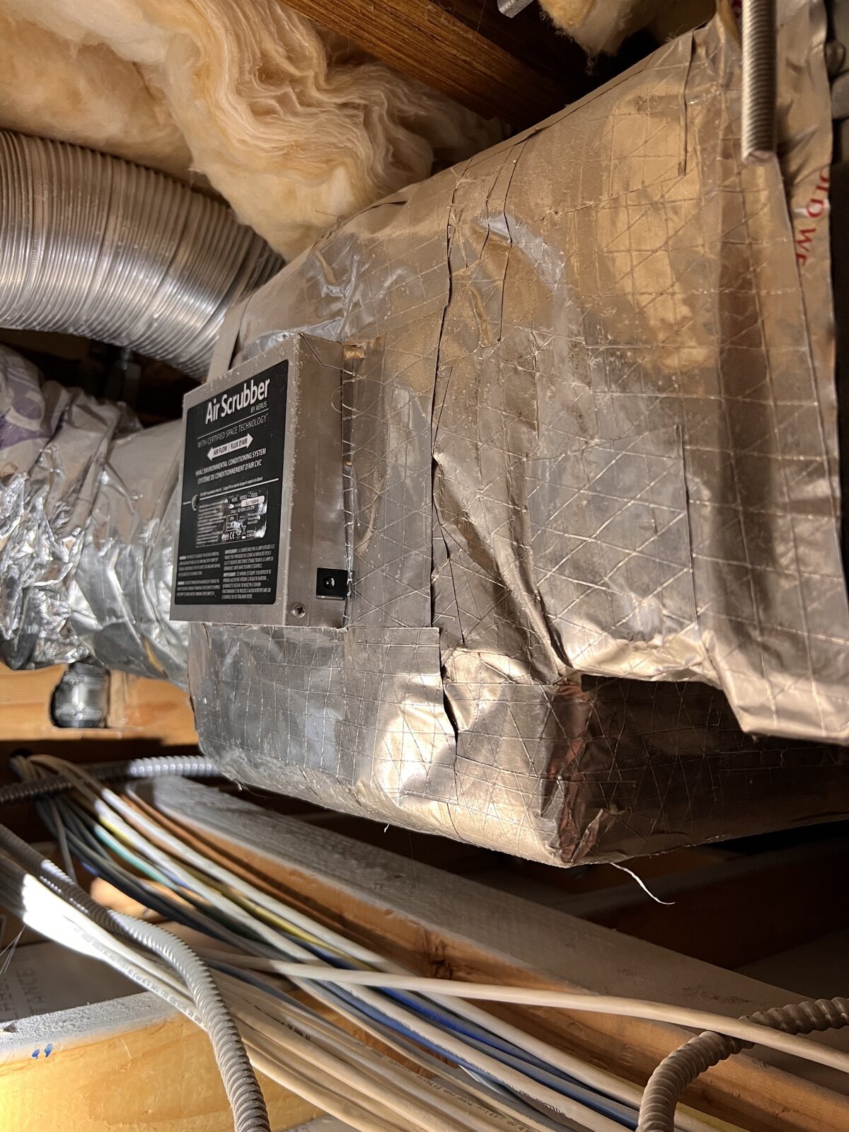 A foil-wrapped duct section in an attic, with a device mounted on its side.