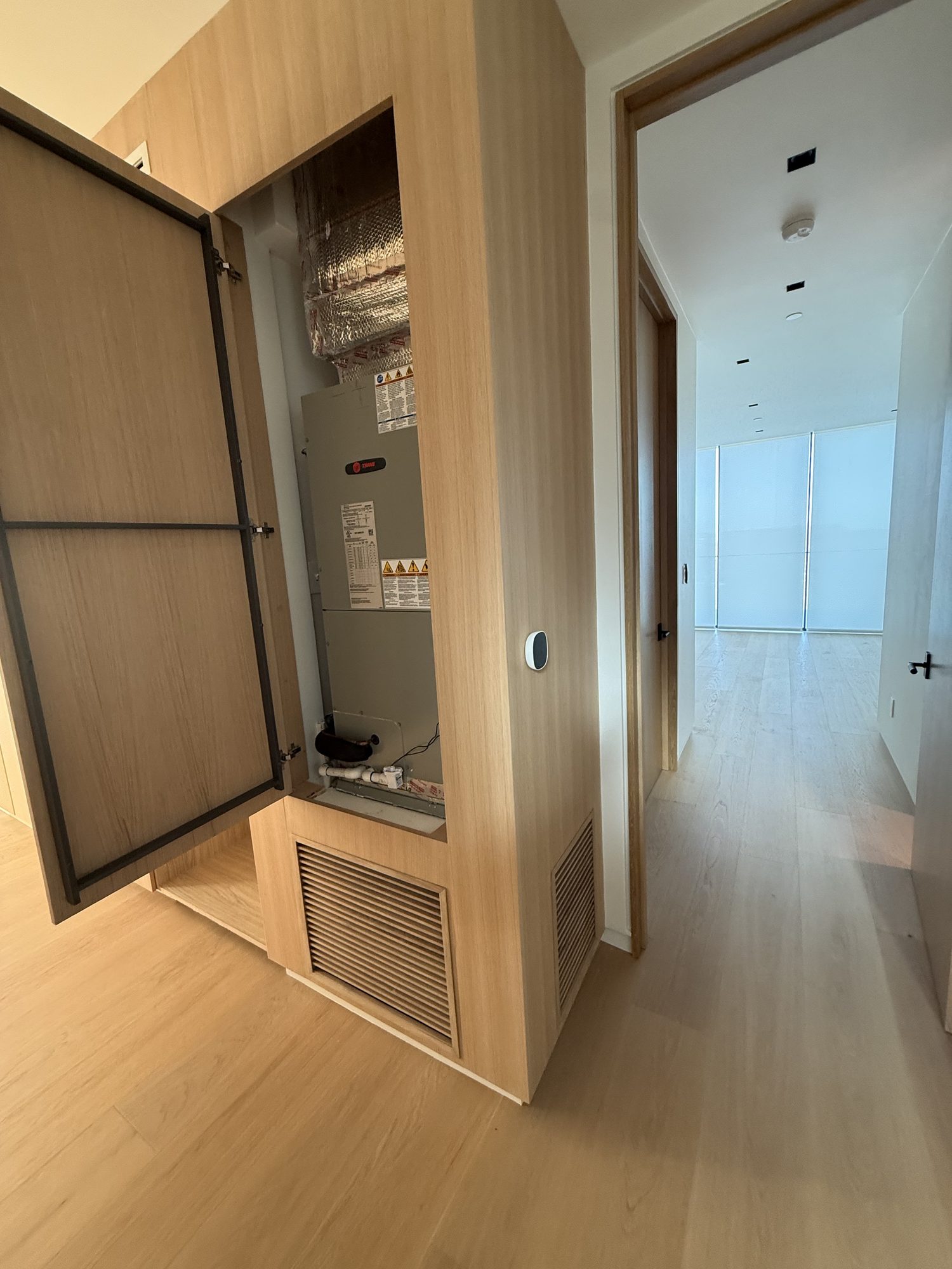 An air handler inside an open oak cabinet in a hallway, with a thermostat on the wall beside it.